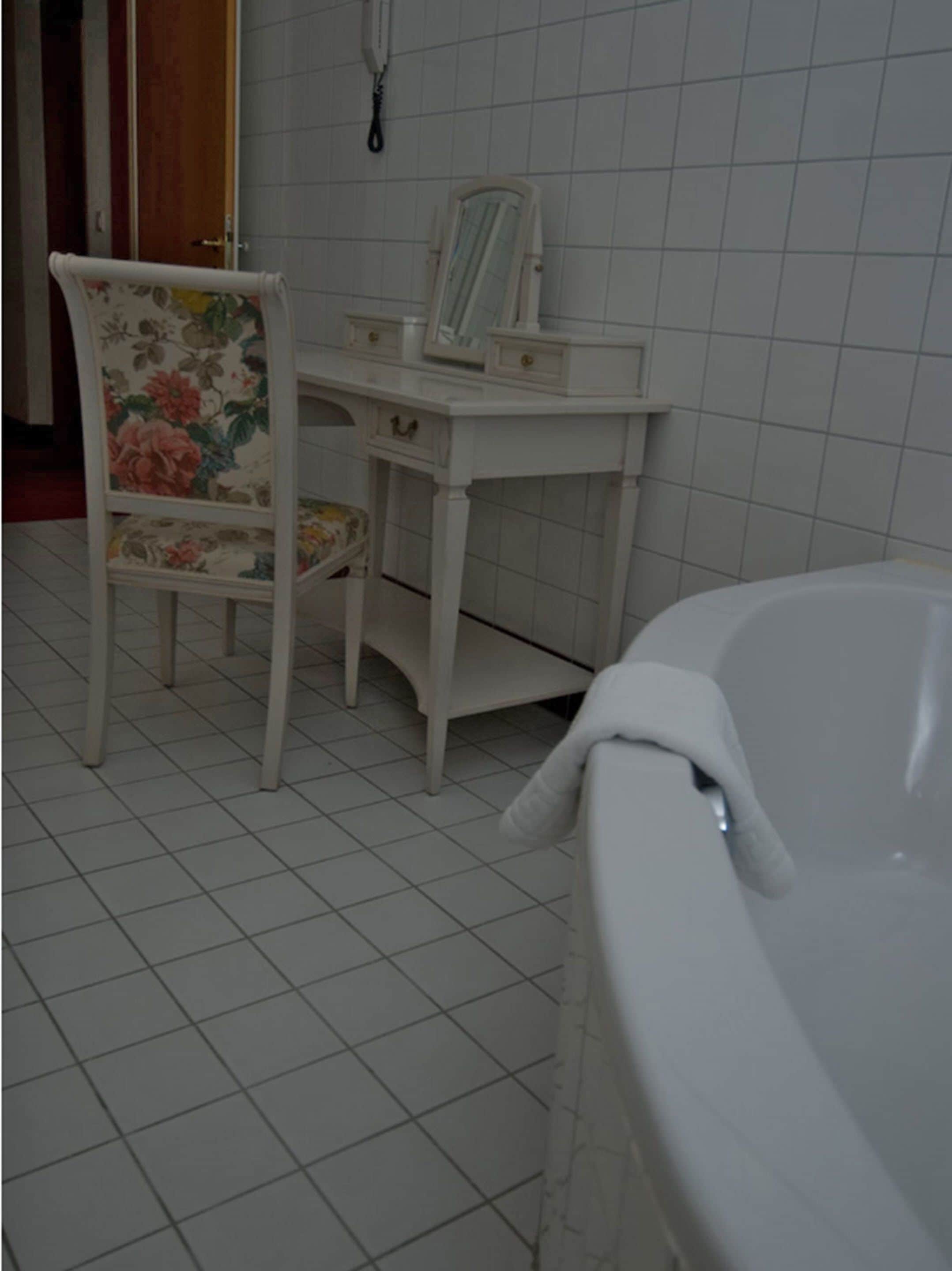 Un baño con una bañera blanca, una silla con tapicería floral, un tocador con espejo y paredes con azulejos.