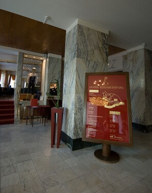 A hotel lobby with marble columns, a reception desk, and a framed signboard detailing hotel amenities.