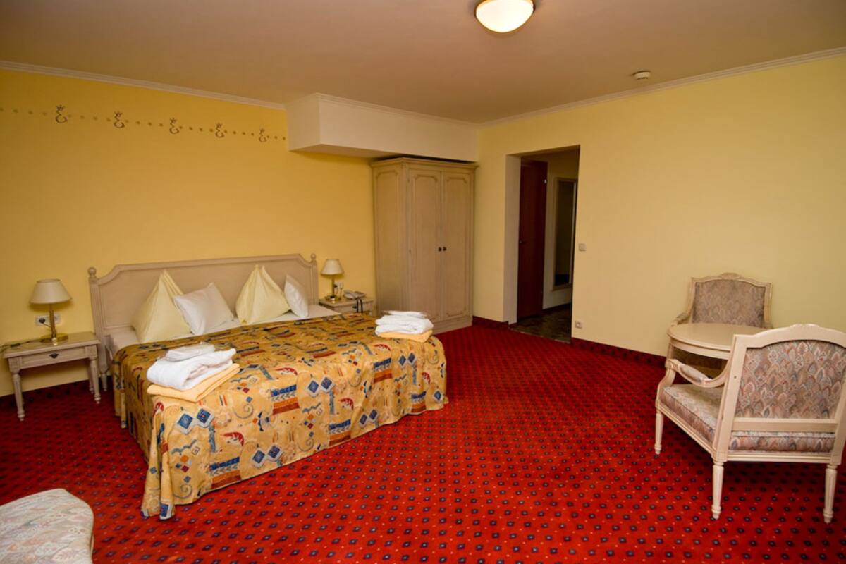 A hotel room with a large bed, a chair, a wardrobe, and a nightstand with a lamp.