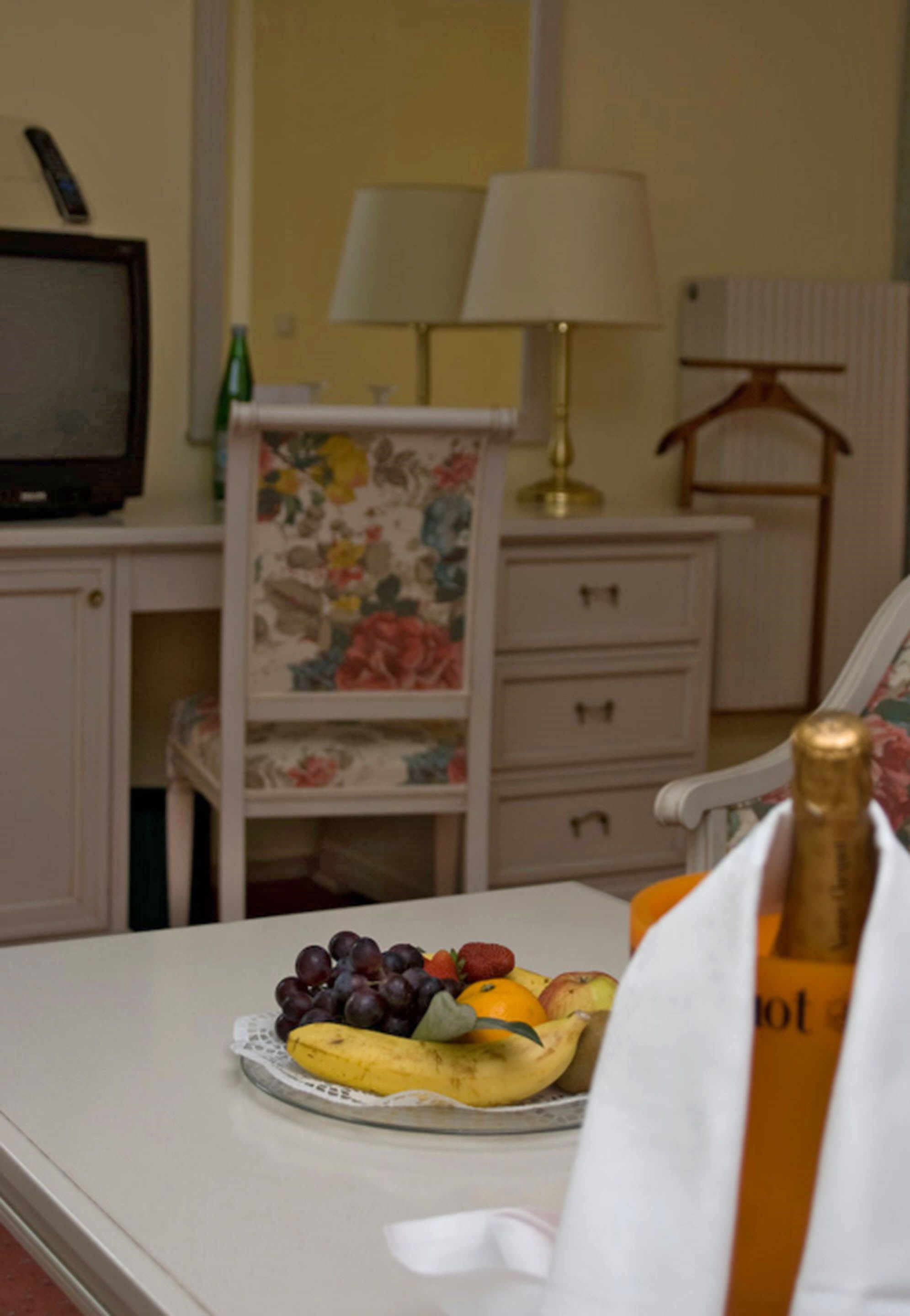 A dining table with a fruit plate, a bottle of champagne, and a television on a stand.