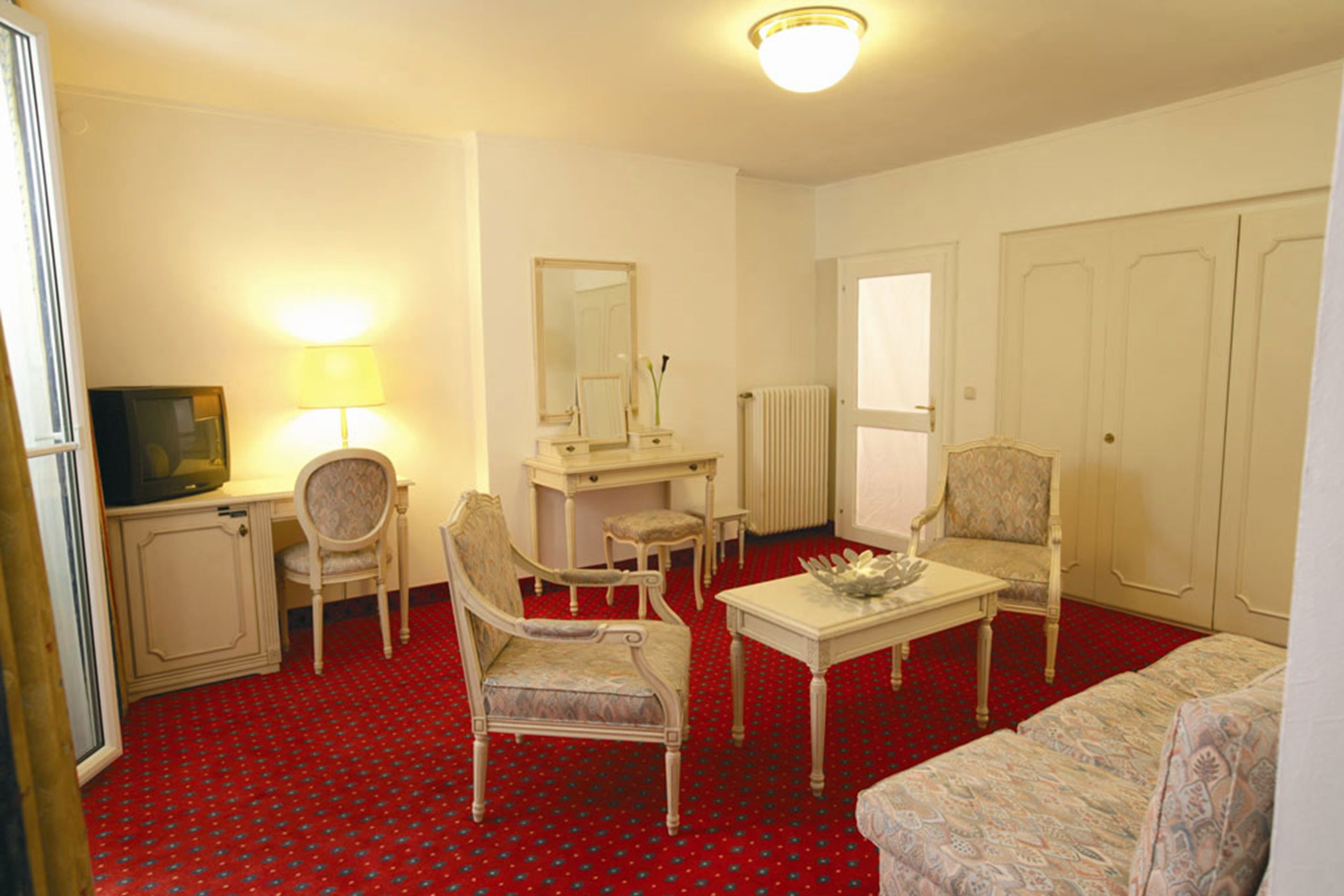 A hotel room with a red carpet, a television, a desk with a lamp, two chairs, a sofa, a mirror, and a radiator.