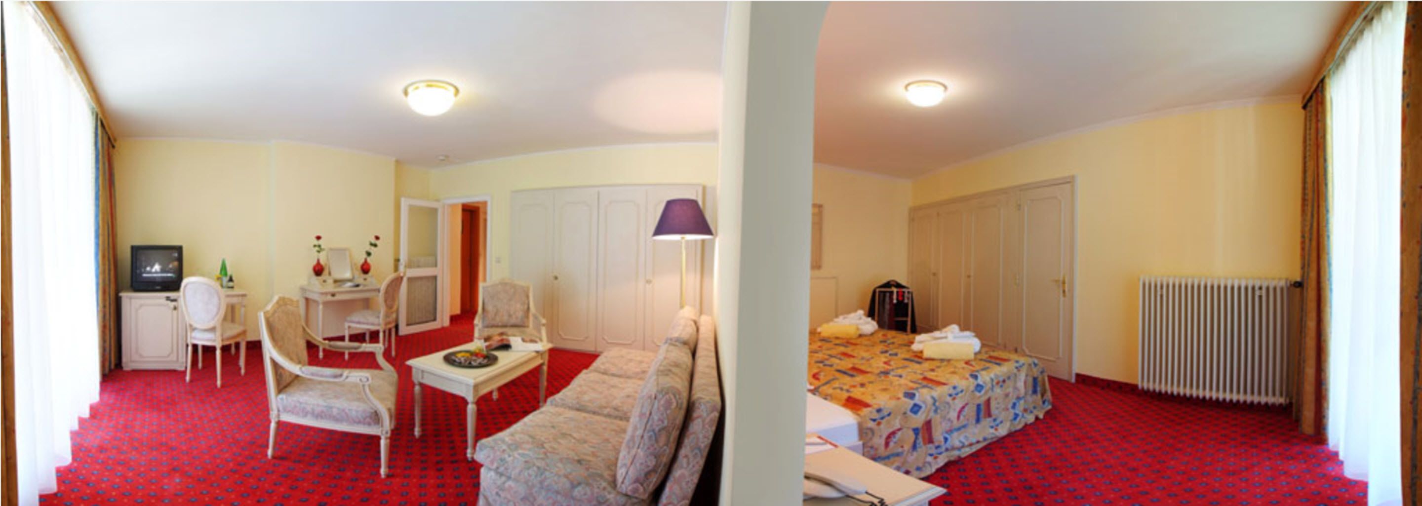Habitación de hotel con alfombra roja, cama con cubrecama estampado, escritorio con silla, televisor y armario.
