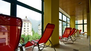Een rij rode lounge fauteuils in een zonnige kamer met grote ramen, een betegelde vloer en uitzicht op een gebouw met een berg op de achtergrond.