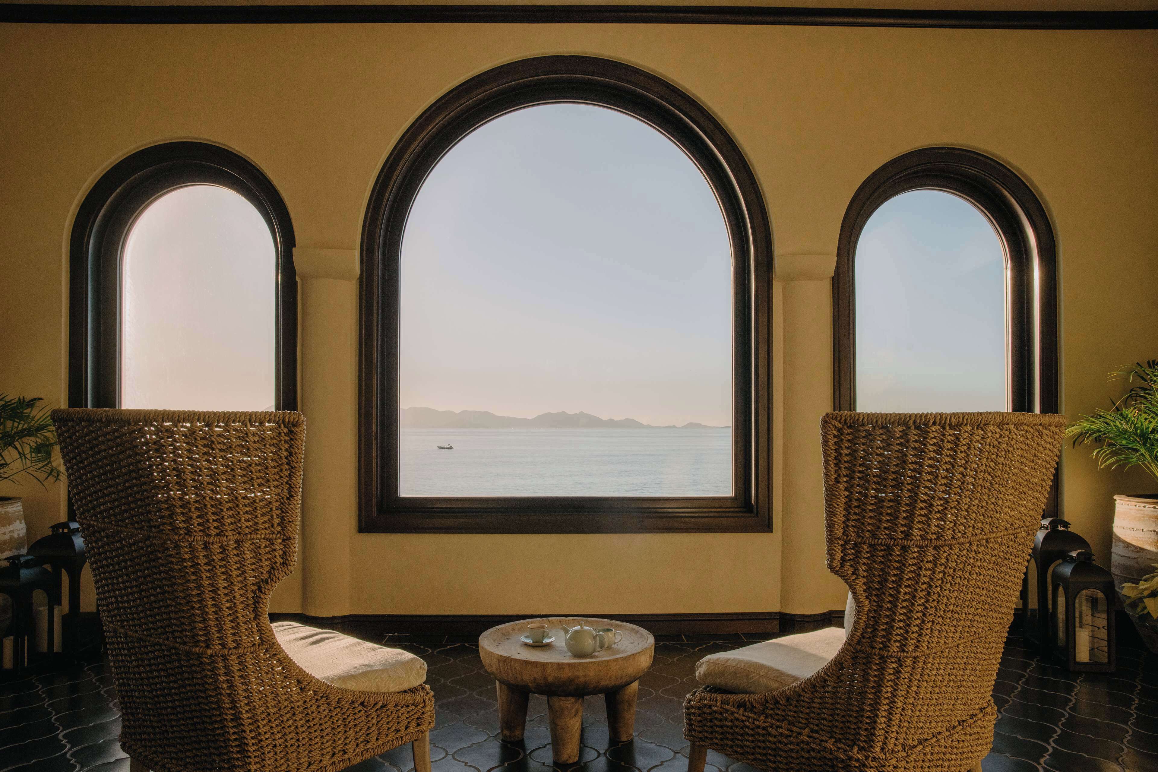 A room with three wicker chairs, a small round table, and a view of the sea through large arched windows.