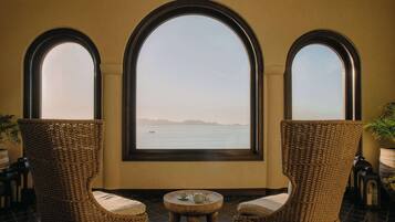 A room with three wicker chairs, a small round table, and a view of the sea through large arched windows.