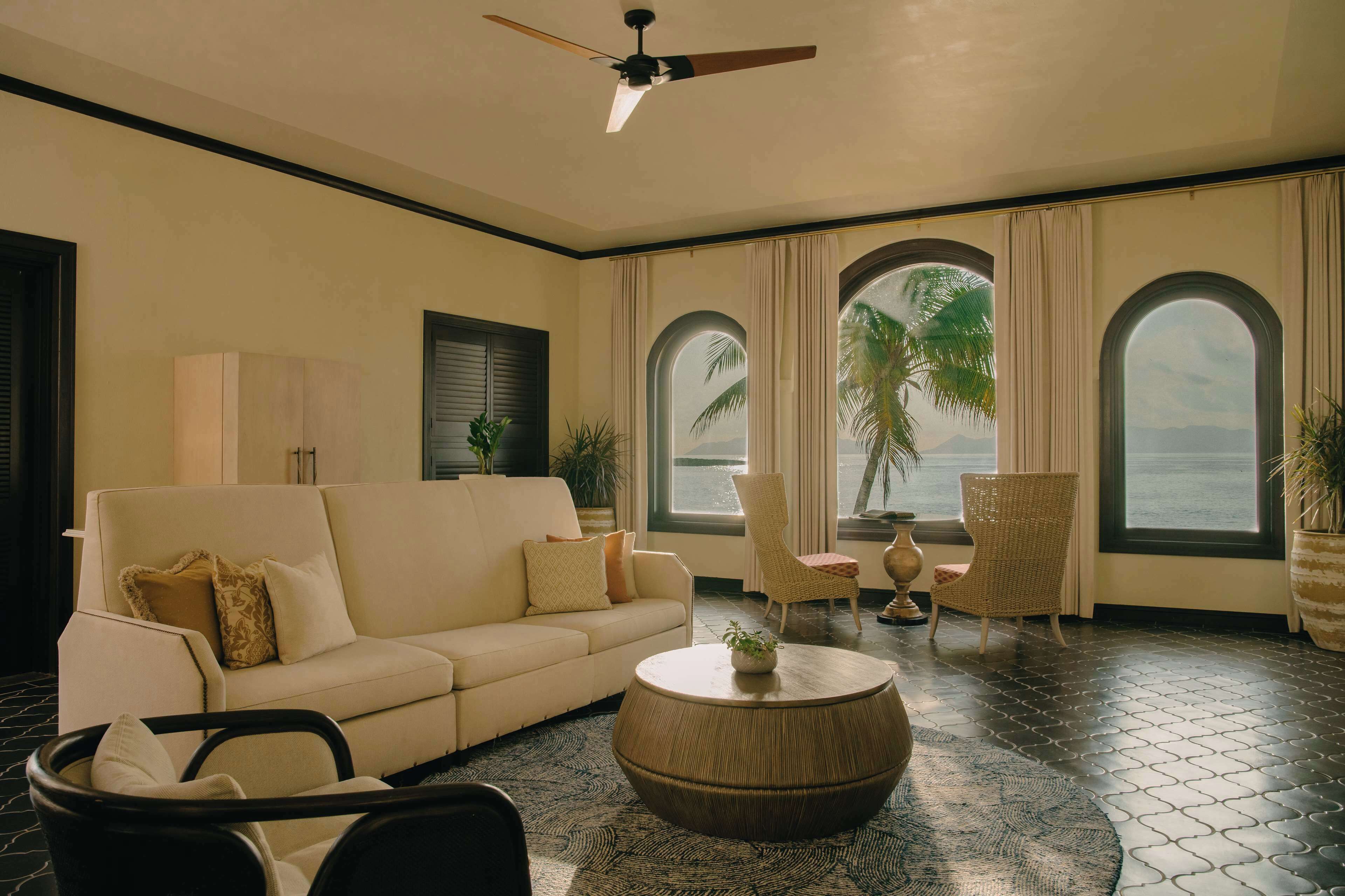 A room with a sofa, chairs, a round coffee table, and large windows with curtains.