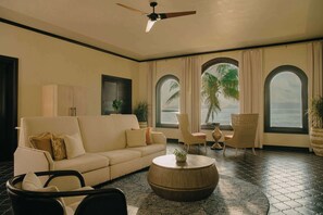 A room with a sofa, chairs, a round coffee table, and large windows with curtains.