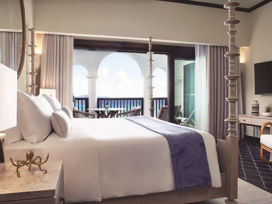A four-poster bed with a balcony view, a desk with a flat-screen TV, and a chair.
