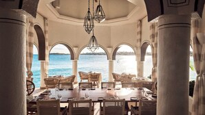 Un espace de restauration intérieur avec vue sur la mer, comprenant une longue table entourée de chaises, des fenêtres en arc de cercle et des luminaires géométriques suspendus.