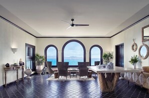 A dining area with a large table, chairs, and a view of the ocean through arched windows.