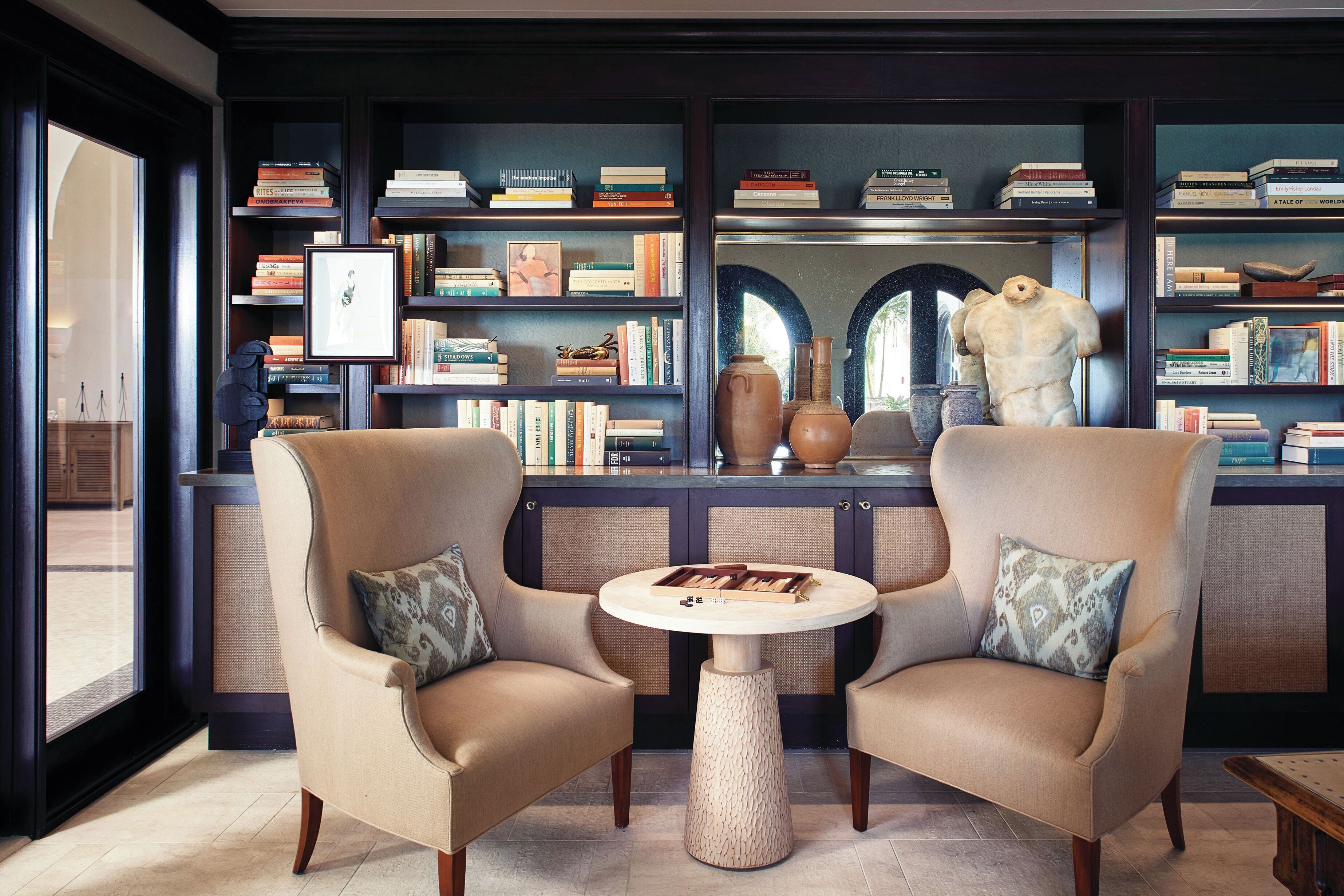 A library with two armchairs, a round table, and bookshelves filled with books and decorative items.