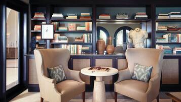 A library with two armchairs, a round table, and bookshelves filled with books and decorative items.