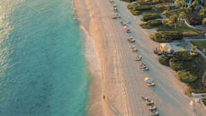 Une vue aérienne d’un complexe hôtelier en bord de mer, avec des rangées de transats et de parasols le long du rivage, entouré de bâtiments blancs et de palmiers.