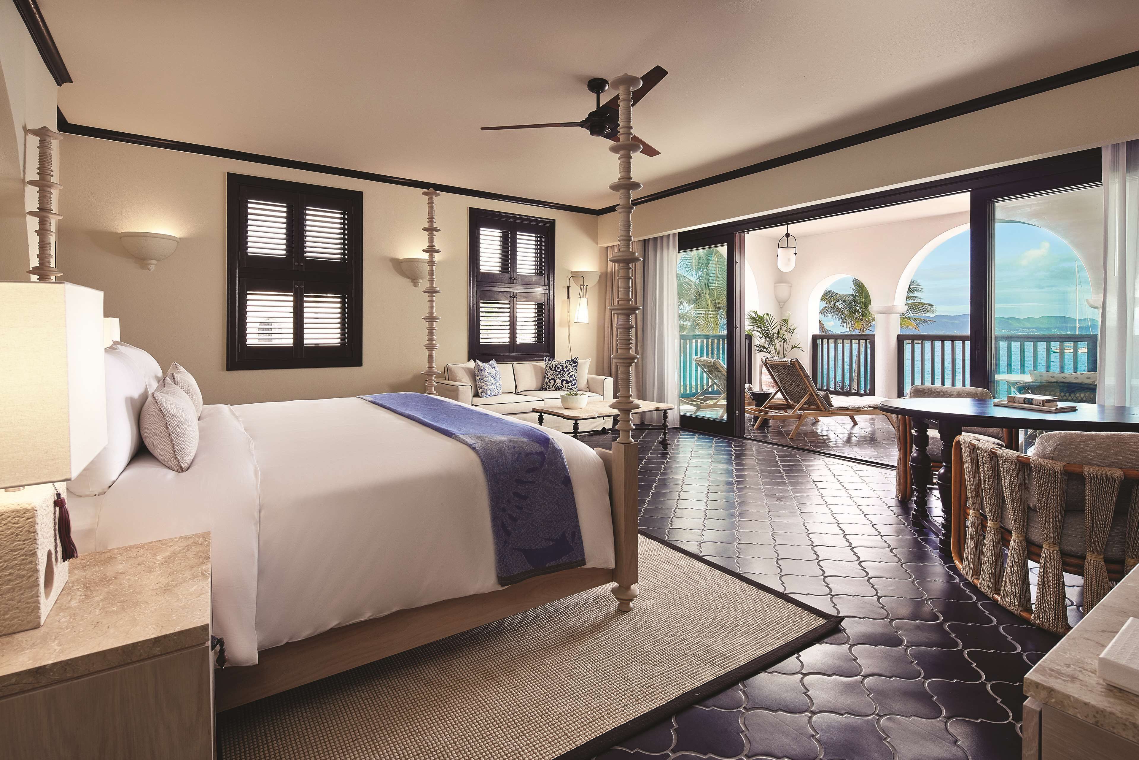 A bedroom with a bed, a ceiling fan, a window with shutters, a balcony with a table and chairs, and a view of the ocean.