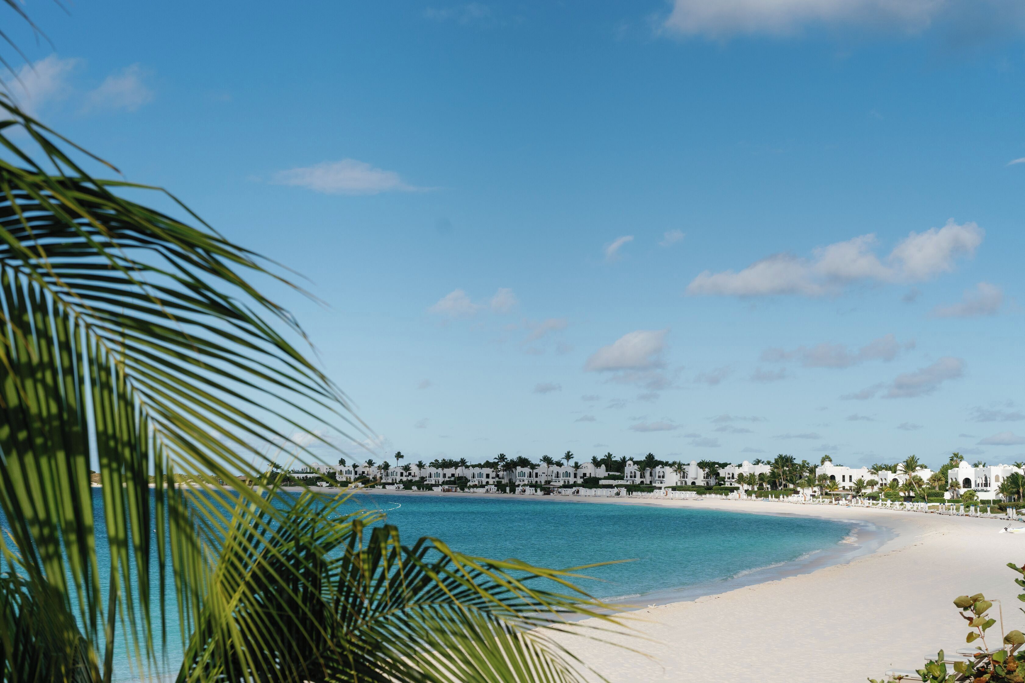 A beach with white sand, clear blue water, and a row of white buildings with palm trees in the background.