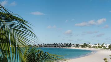 A beach with white sand, clear blue water, and a row of white buildings with palm trees in the background.