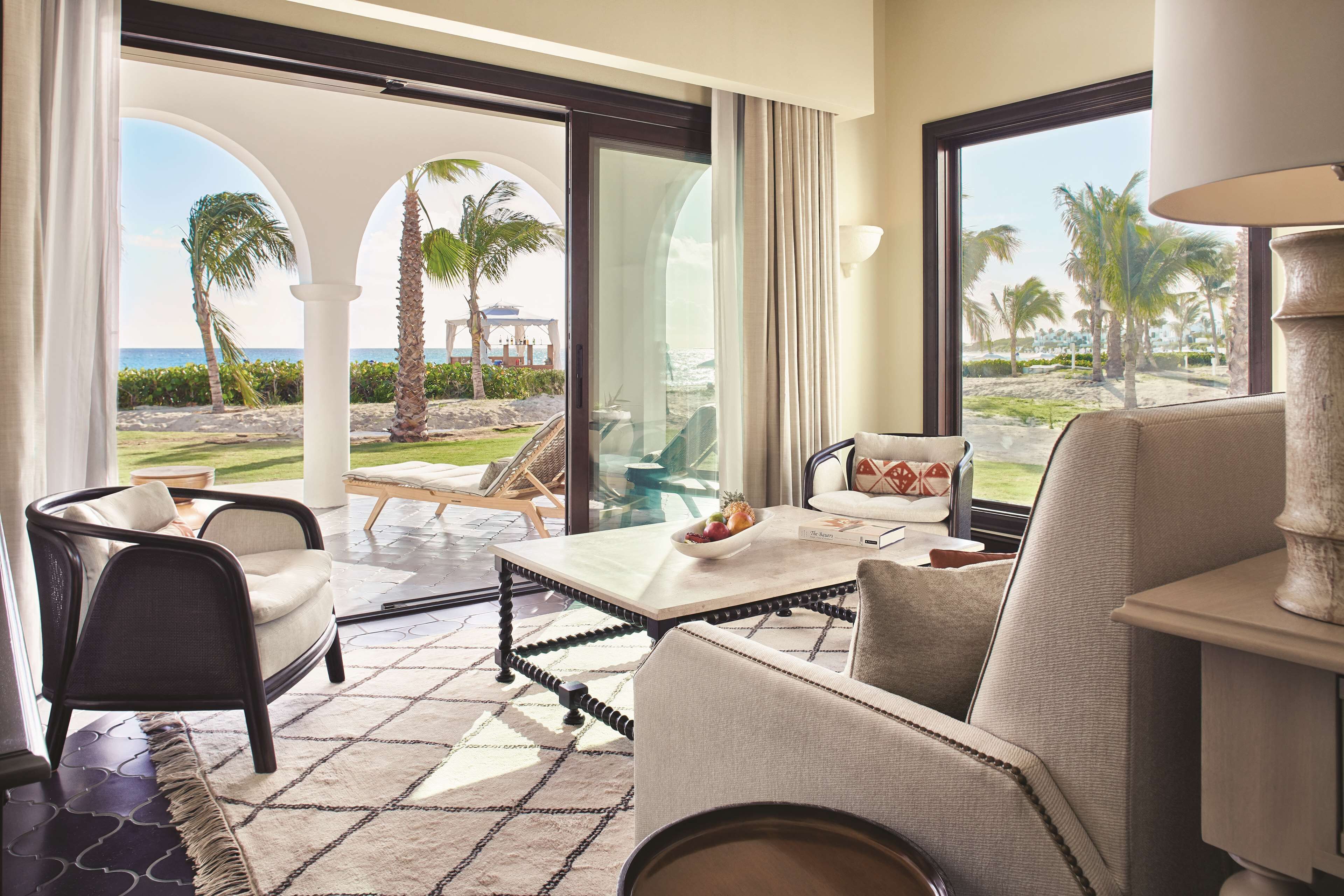 A modern hotel room with a view of the beach, featuring a sofa, armchairs, a dining table, and a large window.