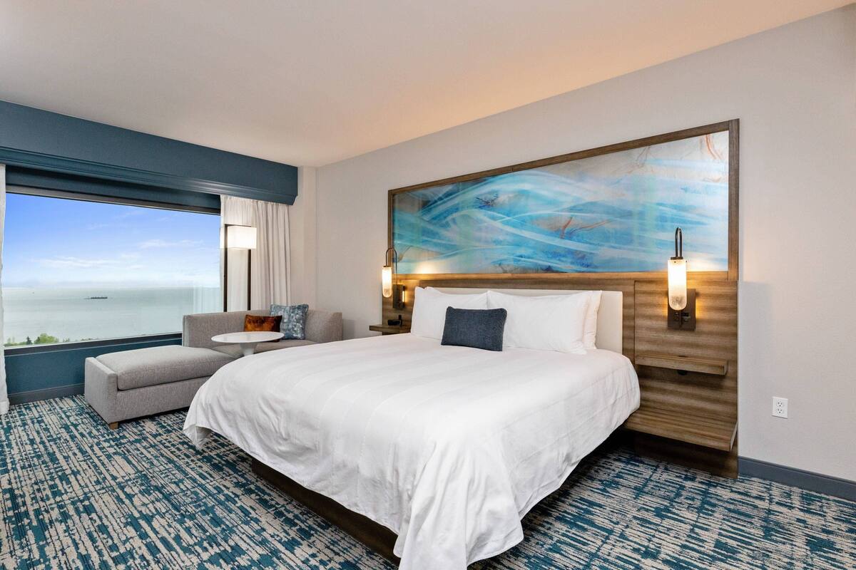 Habitación de hotel con una cama grande, una zona de estar con una mesa, un cuadro en la pared y vista al mar a través de la ventana.