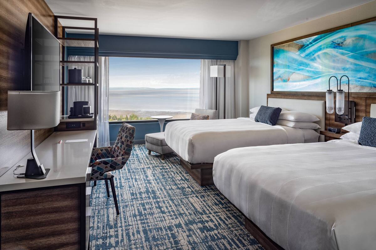 Habitación de hotel con dos camas, un escritorio con silla, televisión y un ventanal con vistas.