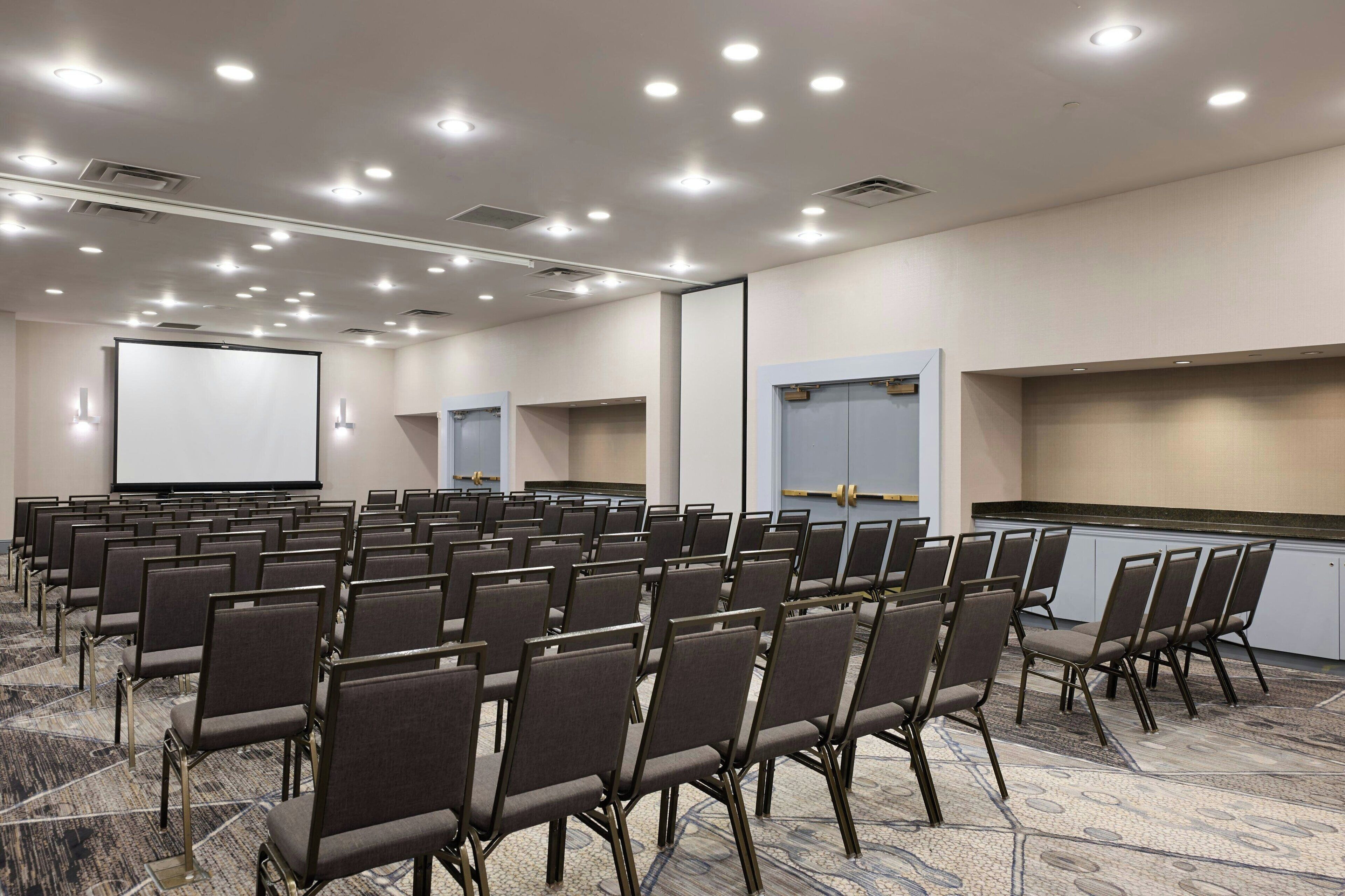 A conference room with rows of chairs, a projector screen, and a large screen on the wall.