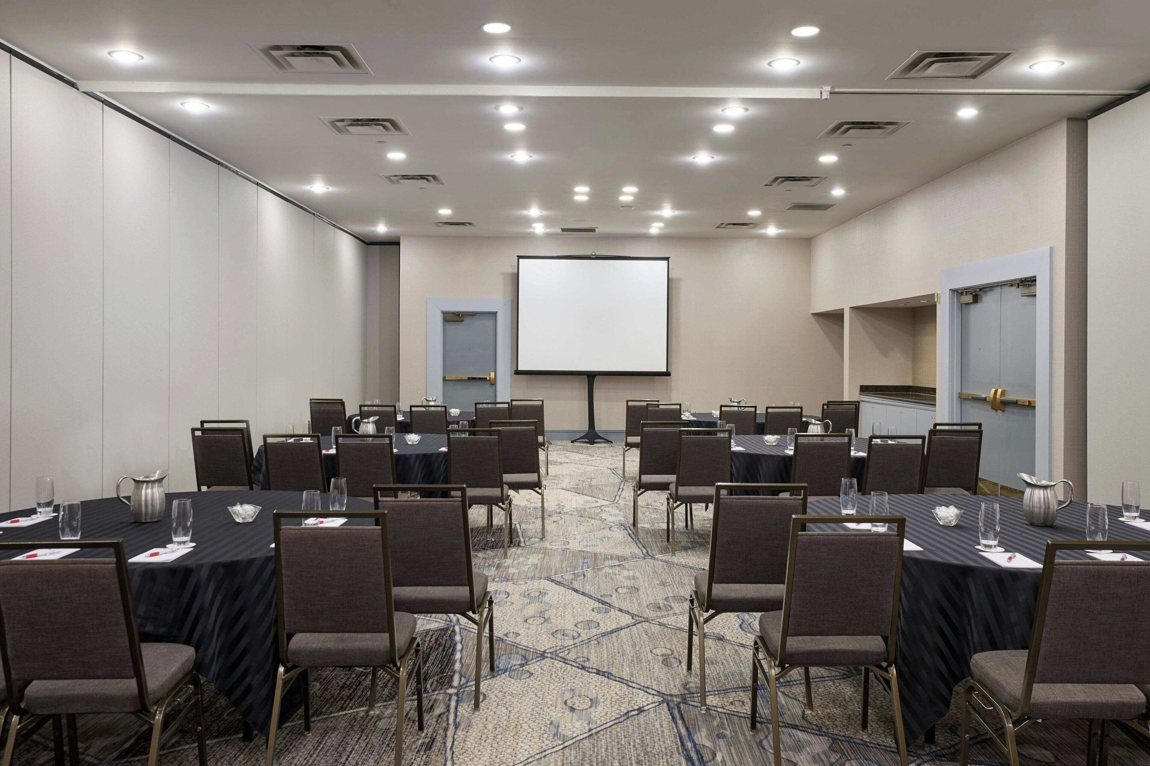 A conference room with multiple tables and chairs, a projector screen, and a carpeted floor.