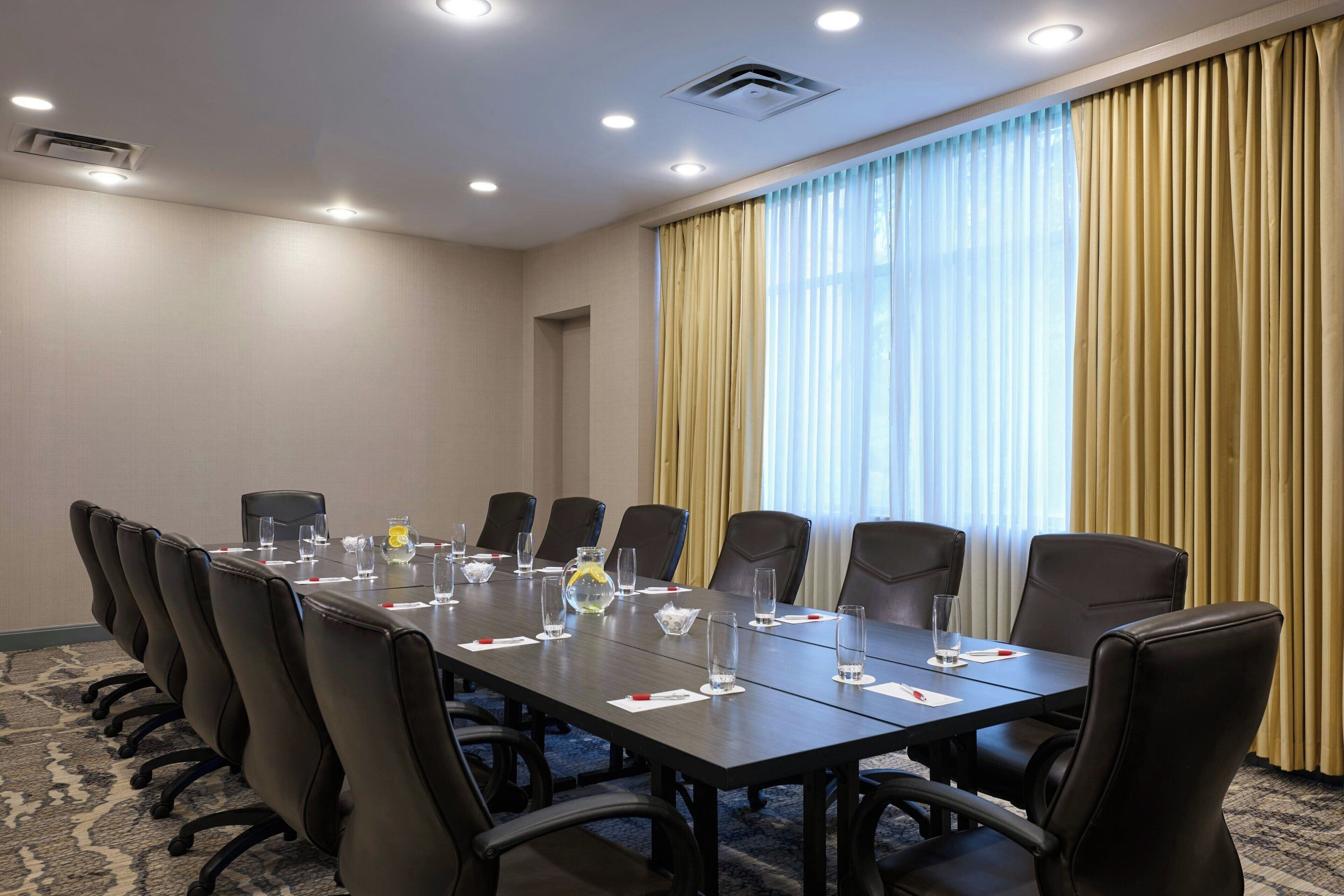 Un salón de conferencias con una mesa larga, sillas negras y un amplio ventanal con cortinas.