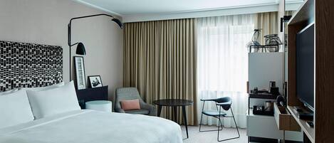 A hotel room with a large bed, a desk with a TV, a chair, a small table, and a lamp.