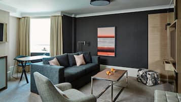 A modern hotel room with a grey sofa, a small table, a chair, and a large window with curtains.