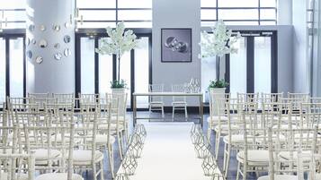 A modern, well-lit room with white chairs arranged in rows, a central table, and large windows providing natural light.