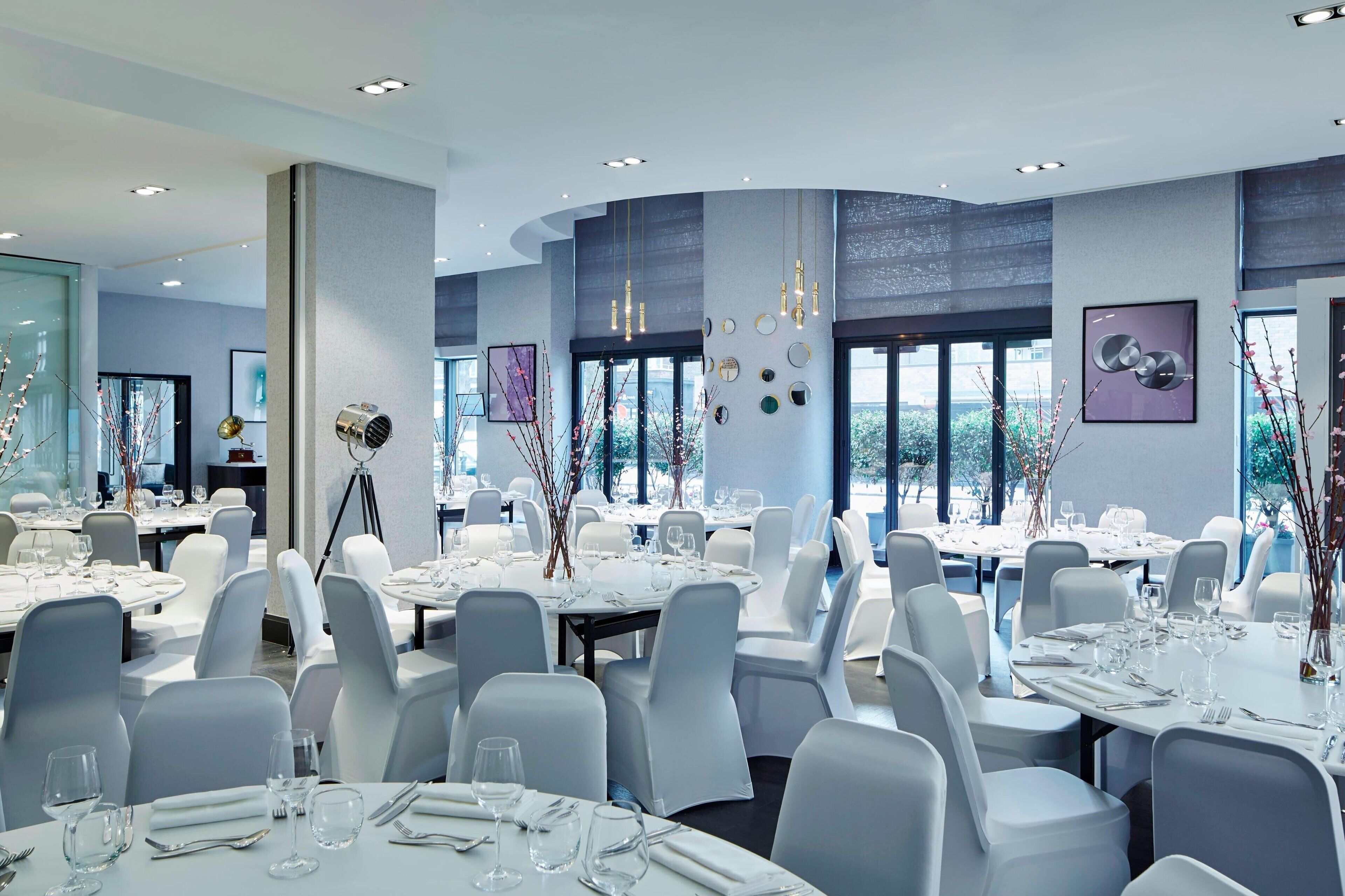 A modern dining area with round tables, white chairs, and a large window providing a view of the outdoors.