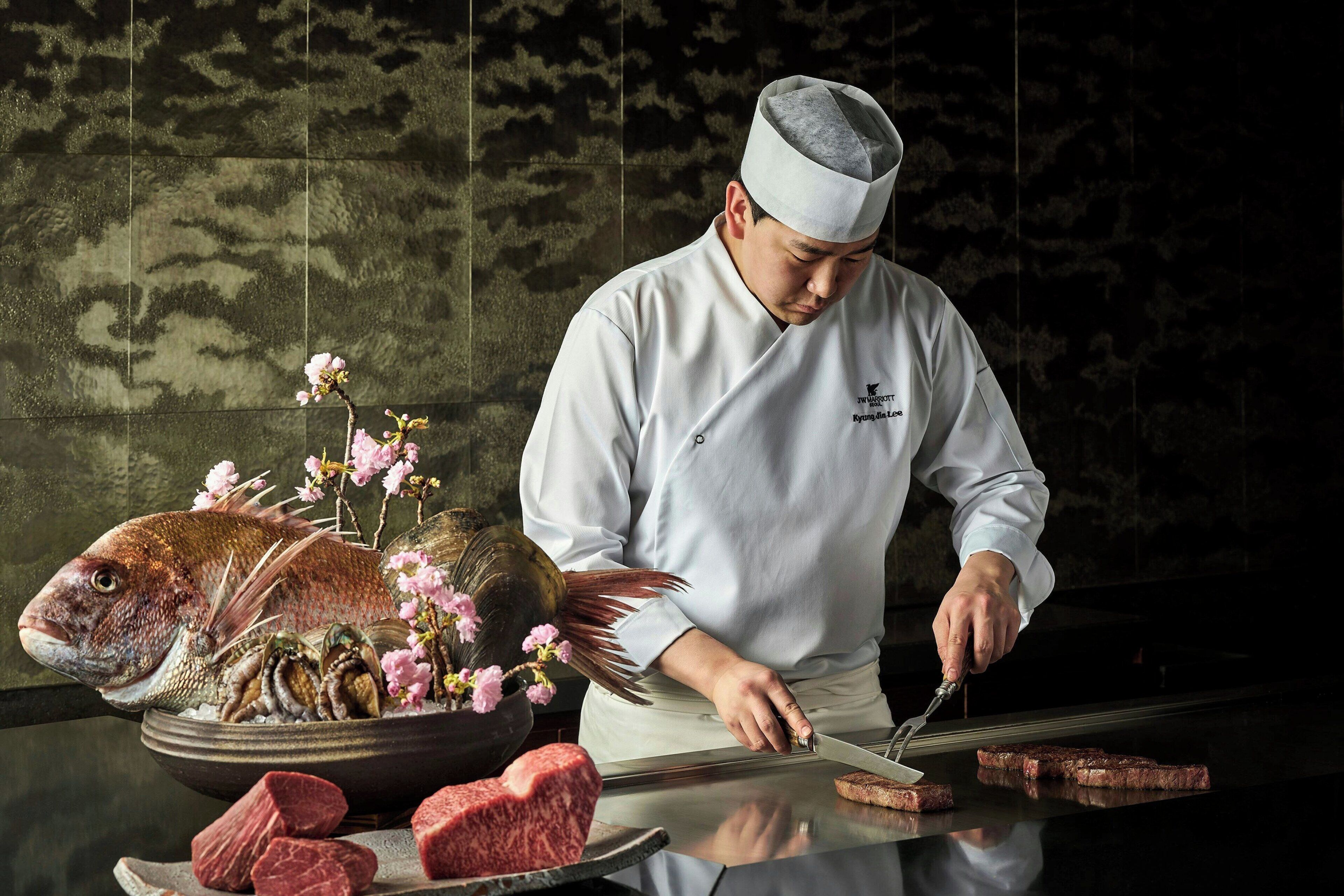 A chef preparing food at a counter with a fish, flowers, and meat.