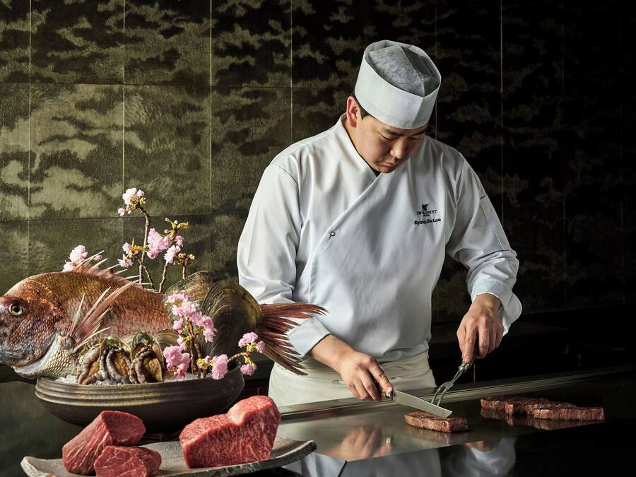 A chef preparing food at a counter with a fish, flowers, and meat.