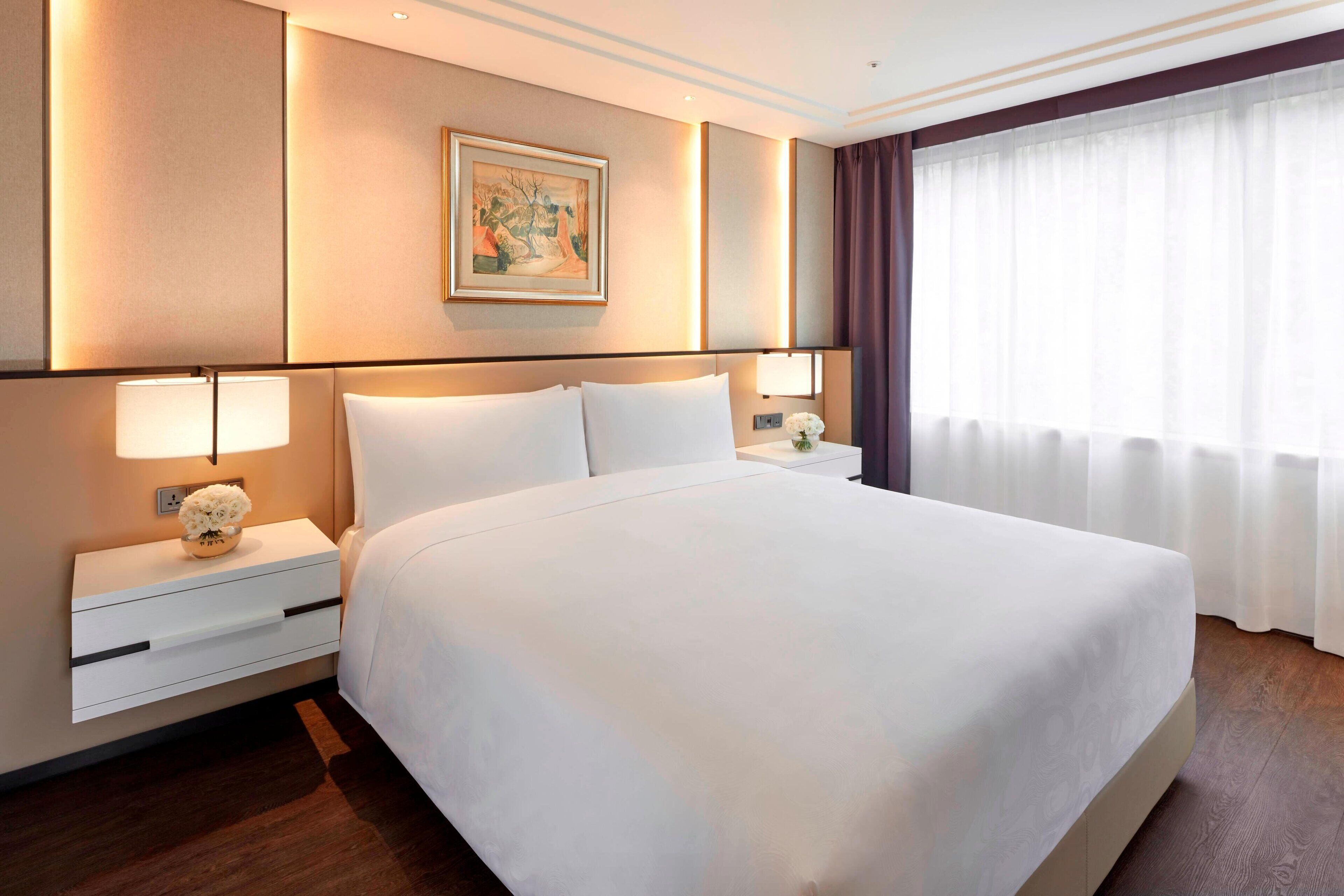 Habitación de hotel con una cama grande, dos mesitas de noche con lámparas, un cuadro en la pared y una ventana con cortinas.