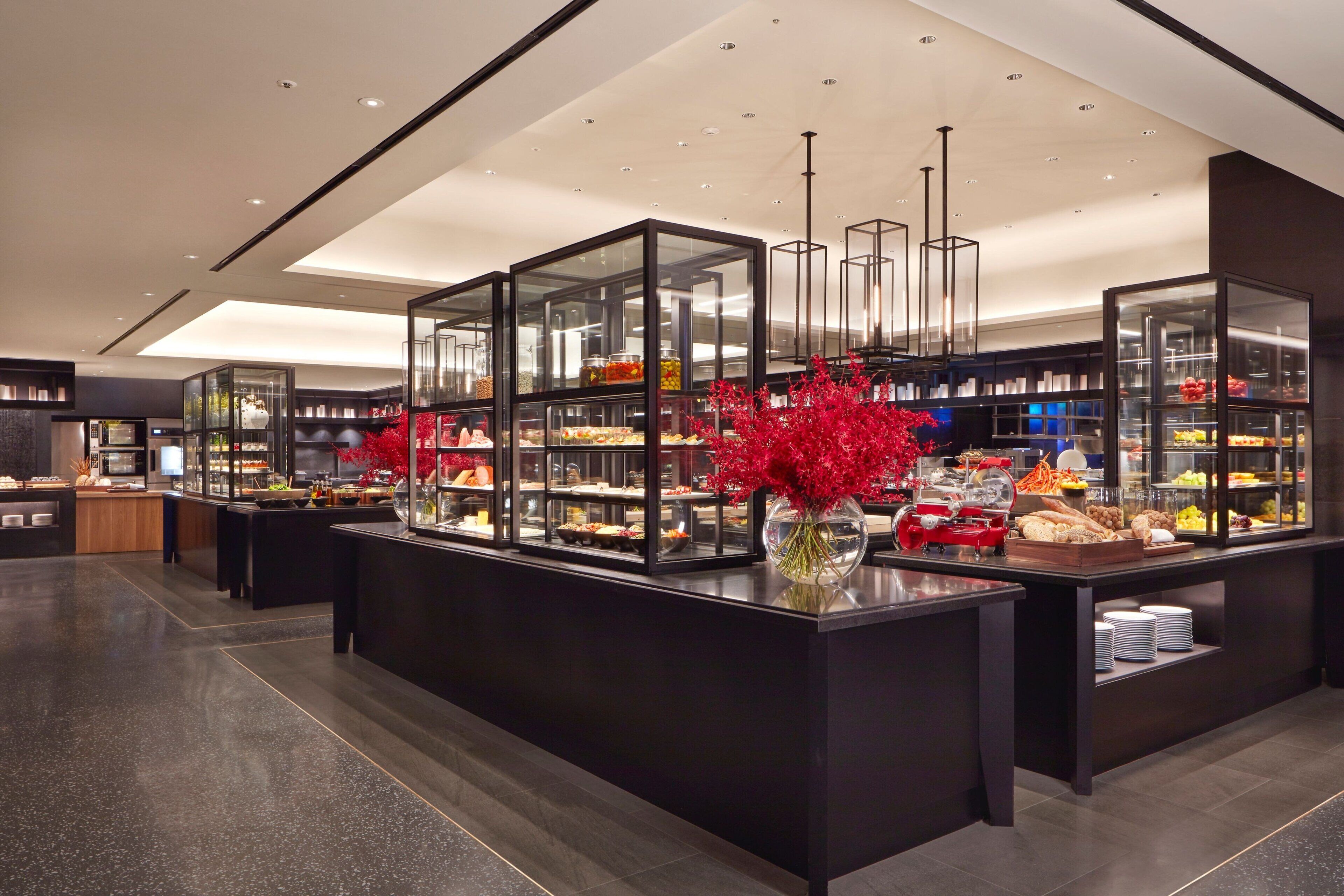 Um saguão de hotel moderno com uma área de buffet central, com vitrines de vidro repletas de diversos alimentos e um arranjo decorativo de flores vermelhas.