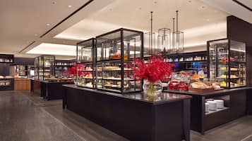 Um saguão de hotel moderno com uma área de buffet central, com vitrines de vidro repletas de diversos alimentos e um arranjo decorativo de flores vermelhas.