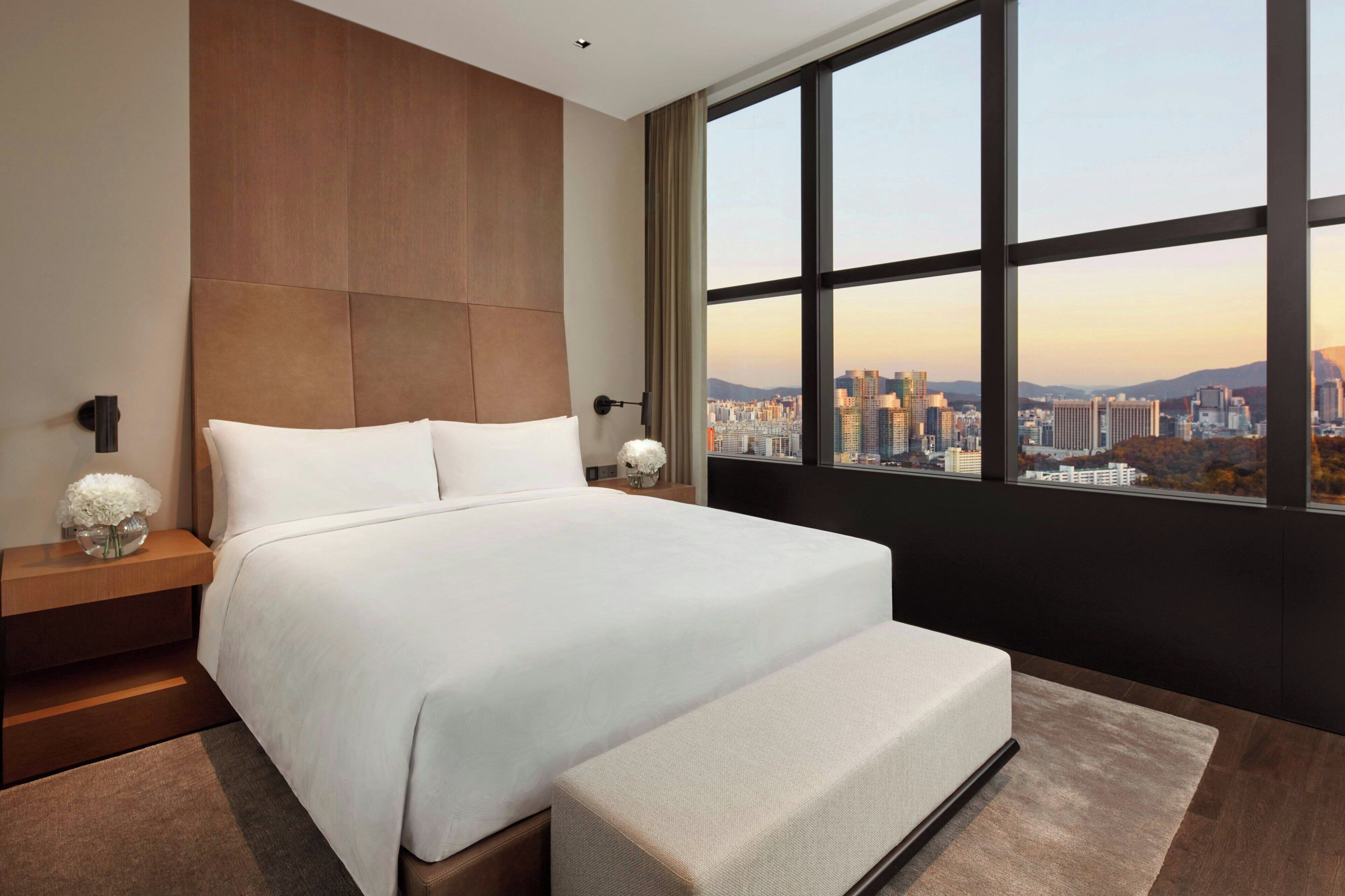 A modern hotel room with a large bed, a nightstand, and a view of a cityscape through a large window.