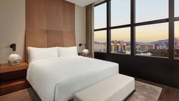 A modern hotel room with a large bed, a nightstand, and a view of a cityscape through a large window.