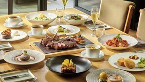 Una mesa de comedor preparada con diversos platos, incluyendo carne, pasta y postres, en una habitación luminosa con ventanas amplias.
