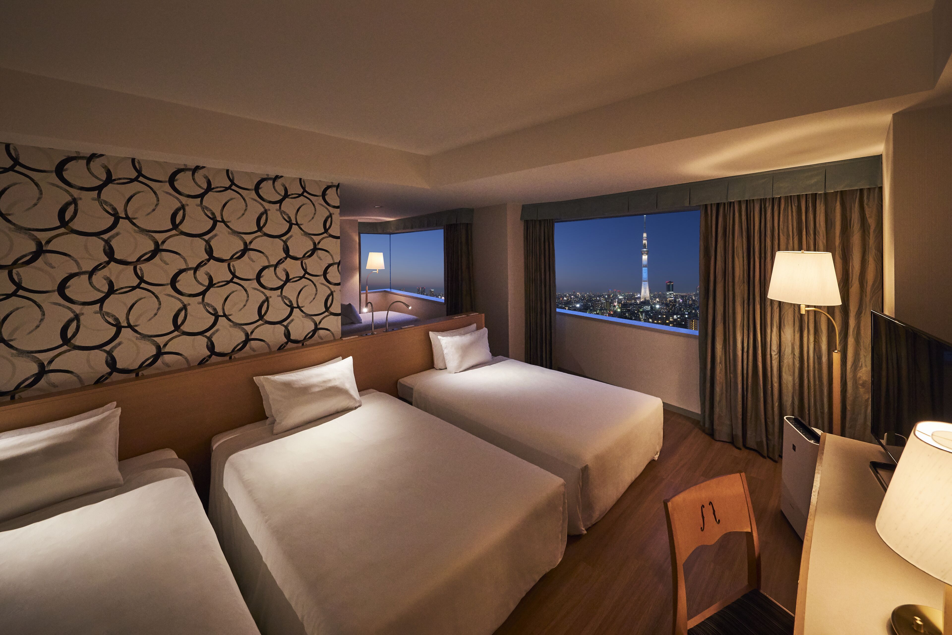A hotel room with two beds, a desk, a chair, a lamp, and a view of a cityscape at night.