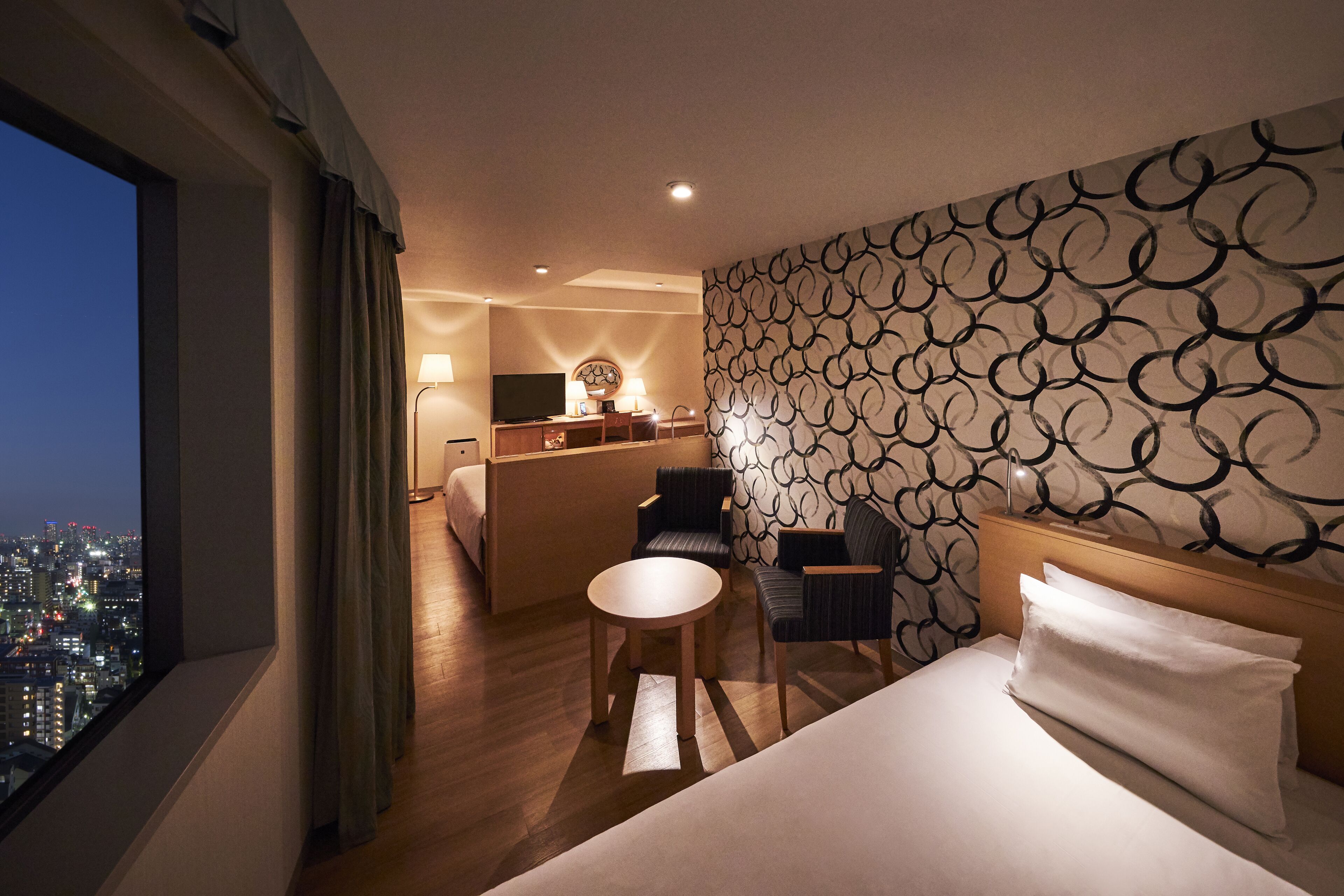 A hotel room with a bed, two chairs, a small table, a desk with a lamp, and a city view.