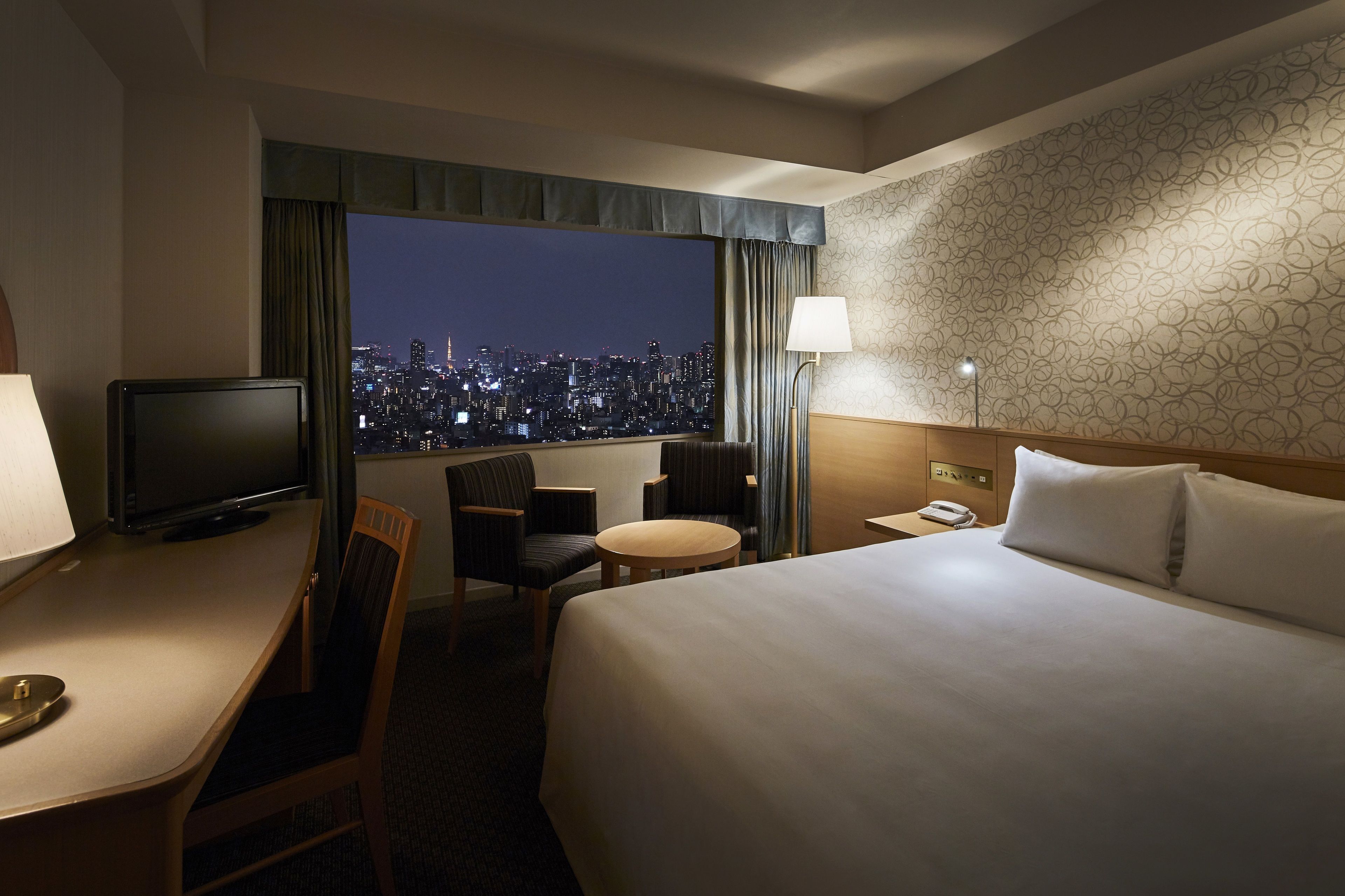 A hotel room with a bed, a desk, a chair, a small table, a television, and a view of a cityscape at night.