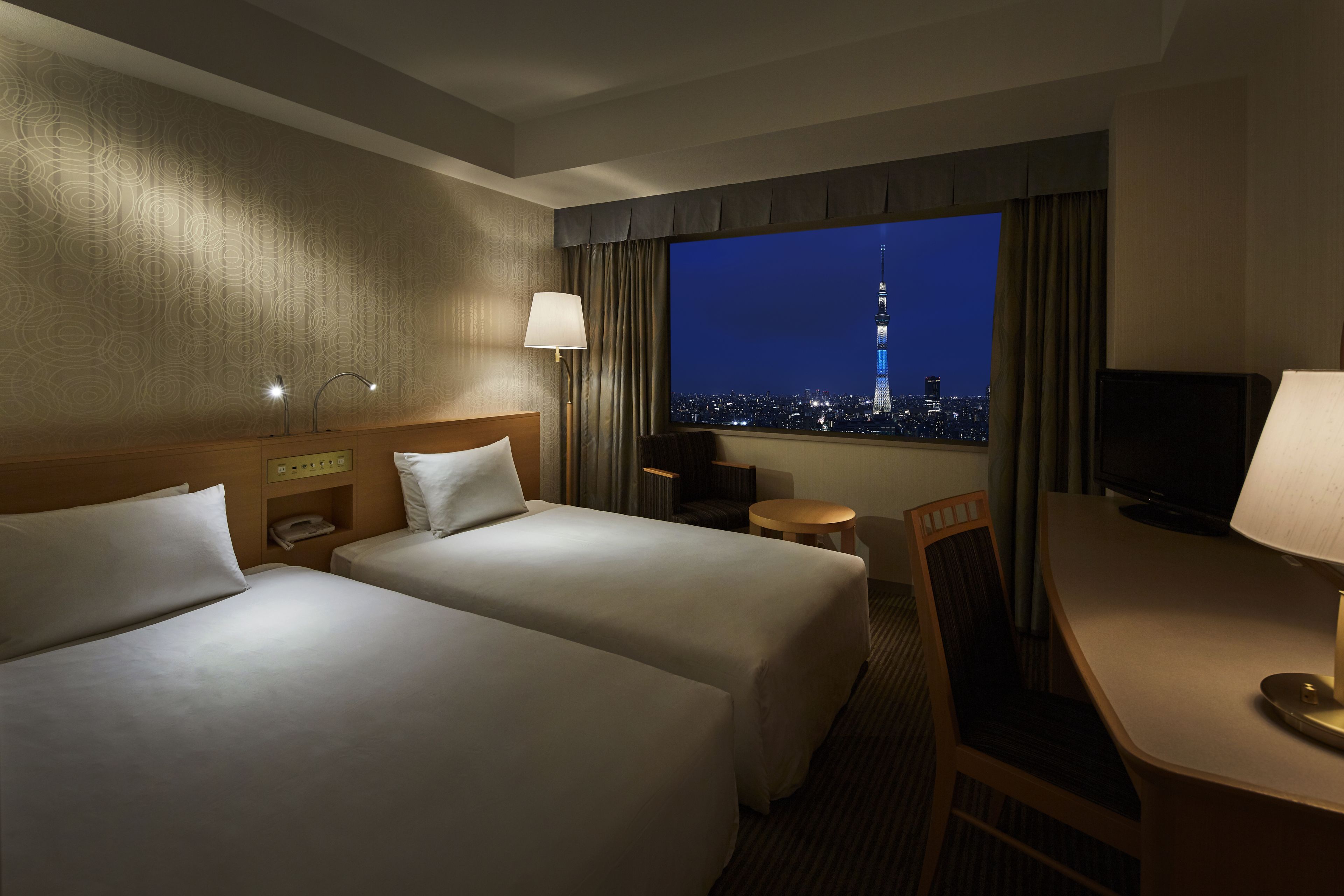 A hotel room with two beds, a desk, a chair, a TV, and a view of a cityscape at night.