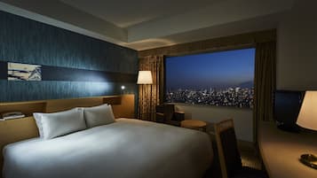 A hotel room with a large bed, a desk with a lamp, a chair, a TV, and a window with a city view.