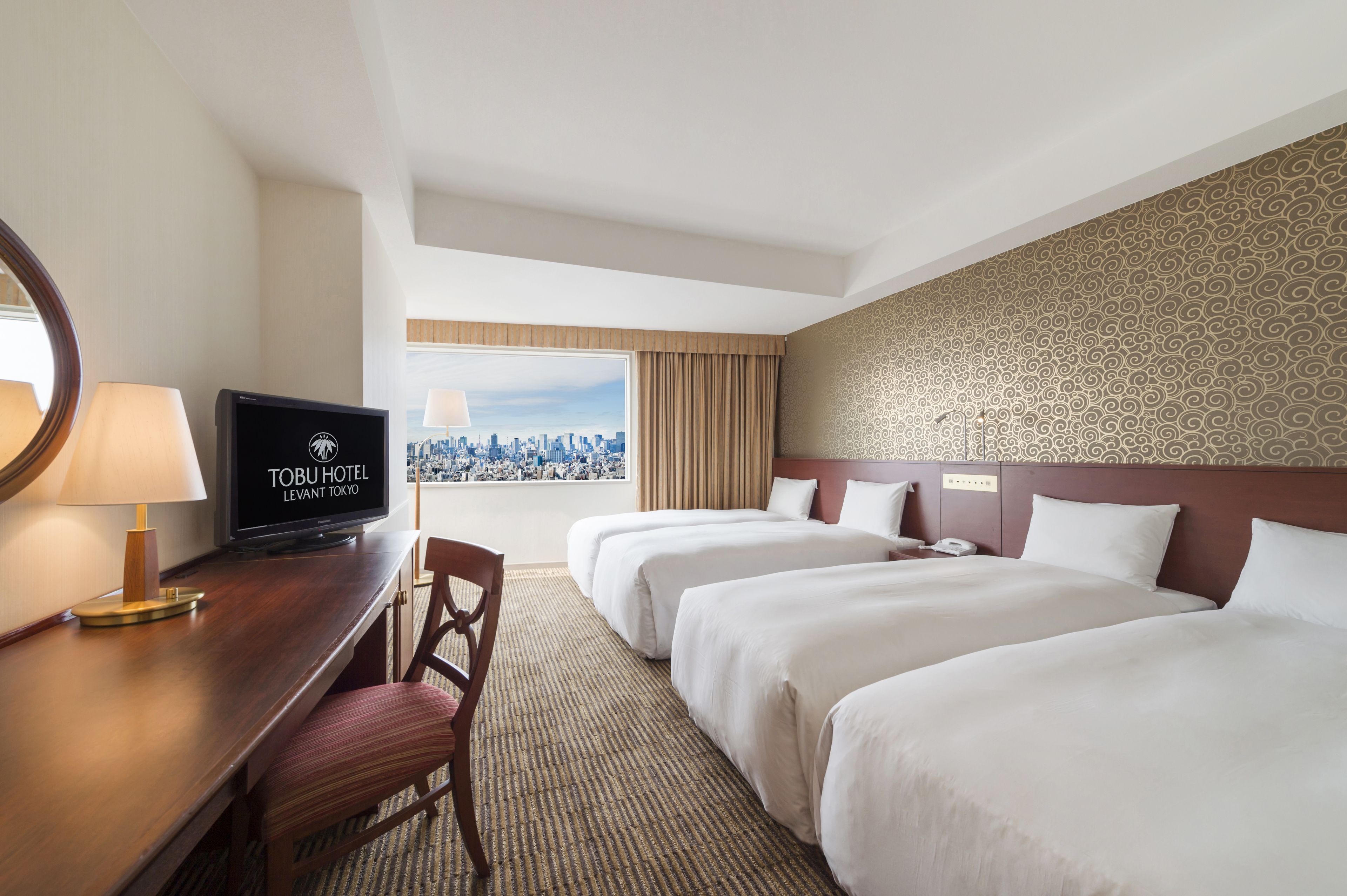 A hotel room with two beds, a desk with a TV, a chair, and a view of the city.