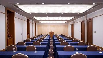 A conference room with rows of tables and chairs, a podium at the front, and a ceiling with recessed lighting.