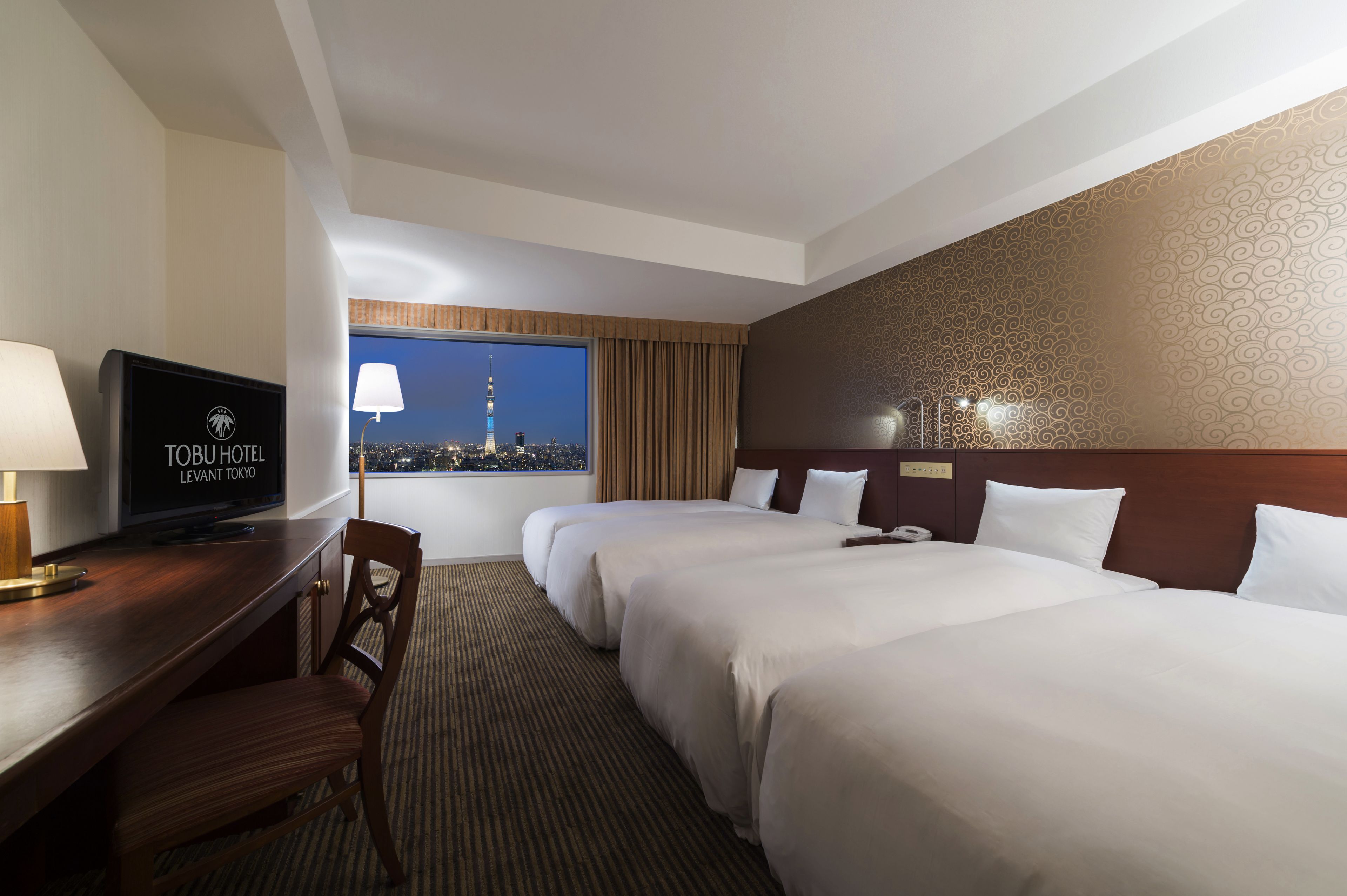 A hotel room with two beds, a desk with a TV, a view of a cityscape, and a patterned wall.