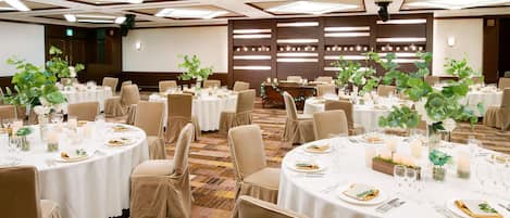Una sala banchetti con tavoli rotondi apparecchiati per la cena, con sedie color beige e tovaglie bianche.