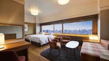 Ein Hotelzimmer mit Bett, Schreibtisch, Stuhl, Sofa und Blick auf eine Stadtkulisse durch große Fenster.