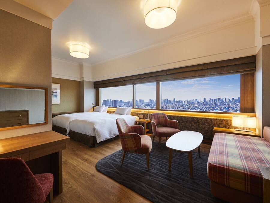 Ein Hotelzimmer mit Bett, Schreibtisch, Stuhl, Sofa und Blick auf eine Stadtkulisse durch große Fenster.