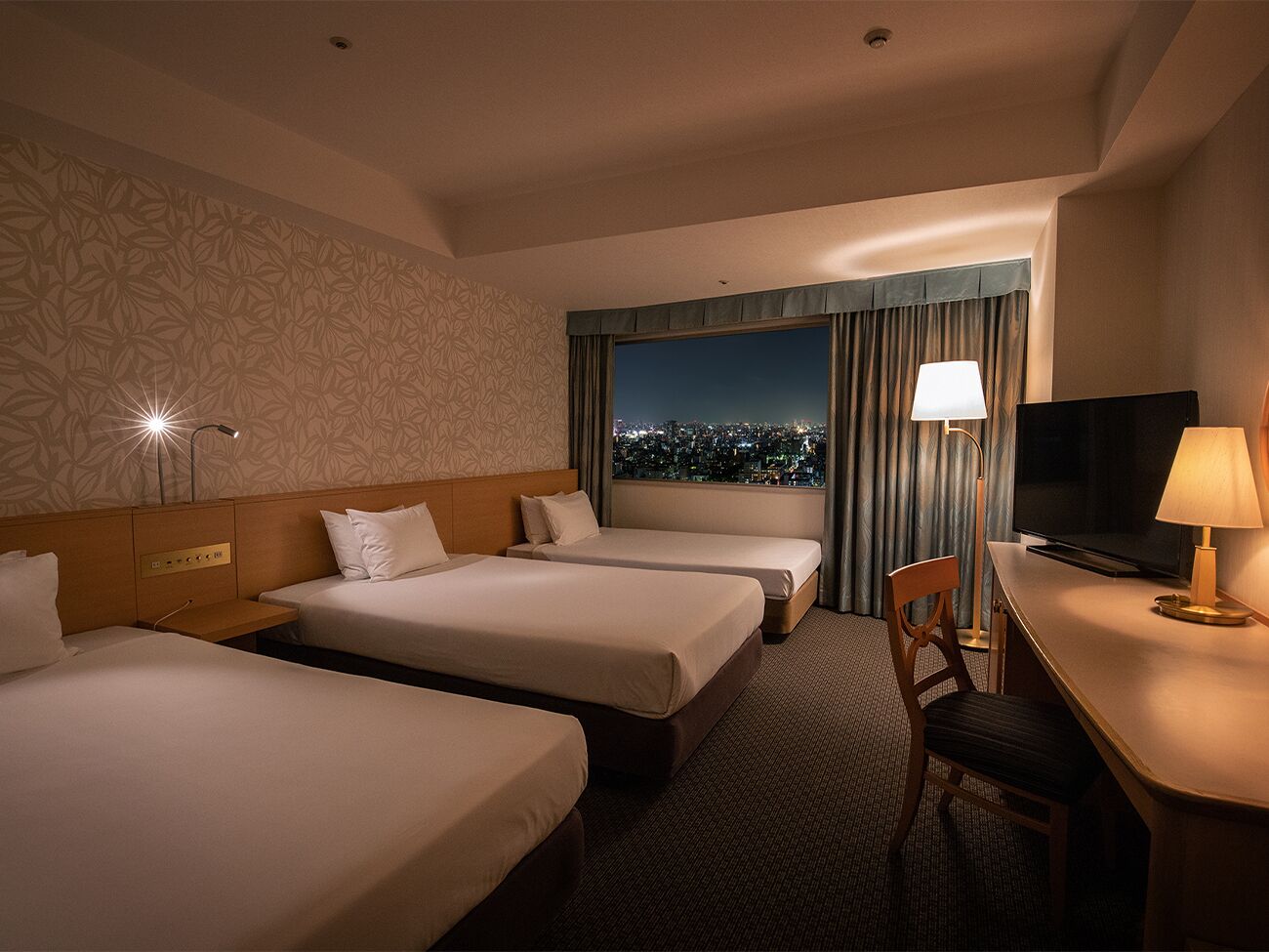 A hotel room with three beds, a desk with a lamp, a TV, and a city view through the window.
